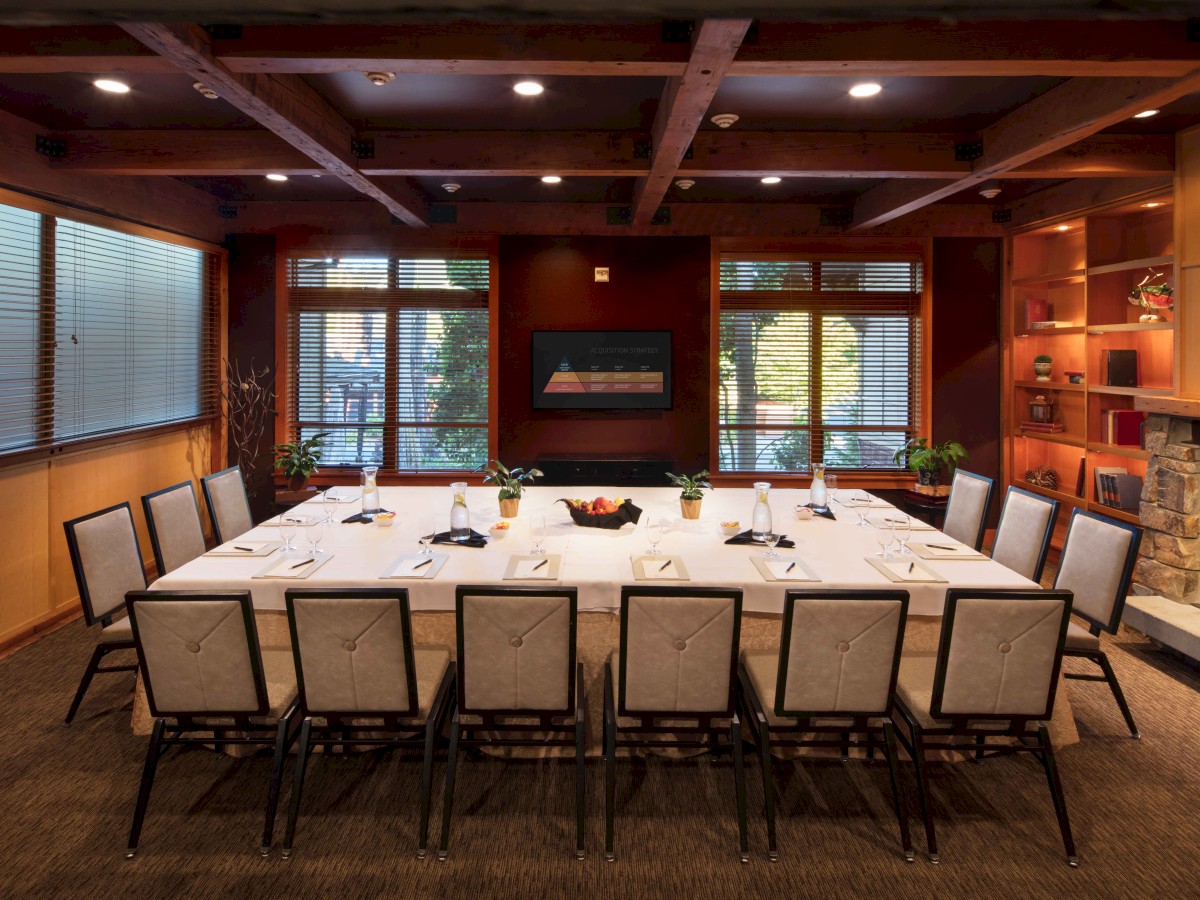 A well-lit conference room with a rectangular table, chairs, a fireplace, bookshelves, and large windows is featured in the image.