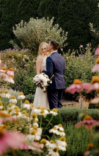 A bride and groom are standing in a garden surrounded by blooming flowers, with the groom in a suit and the bride holding a bouquet of flowers.