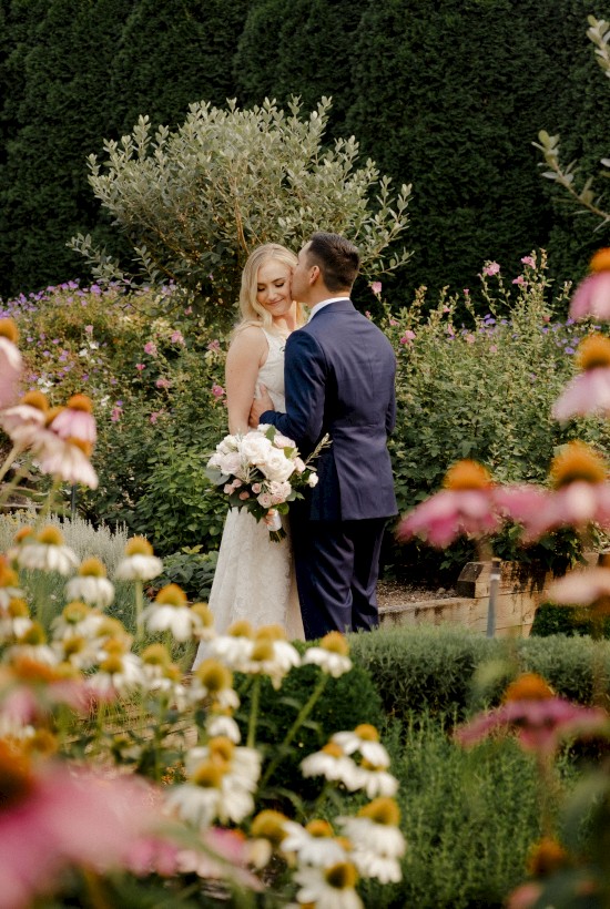 A bride and groom are standing in a garden surrounded by blooming flowers, with the groom in a suit and the bride holding a bouquet of flowers.