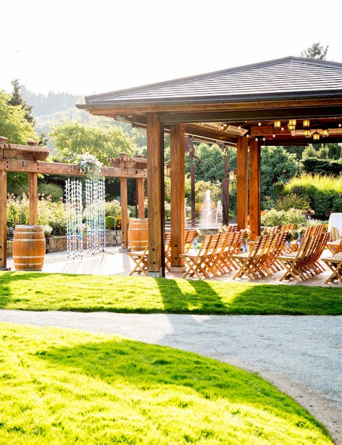 An outdoor venue with a wooden pergola, chairs, barrels, and well-maintained green surroundings in a garden setting.