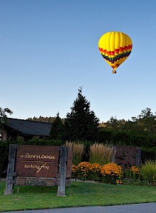 A yellow and red hot air balloon is flying over a landscape with lush greenery, flowers, a building, a sign, and two people walking.