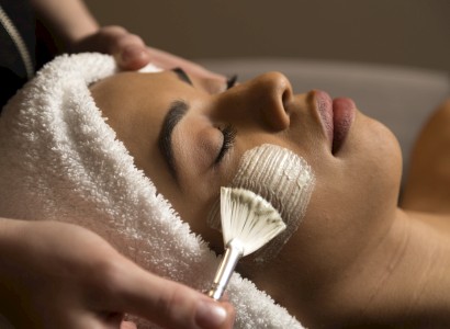 A person is receiving a facial treatment; a brush applies a mask to their cheek while they wear a towel on their head and relax with closed eyes.