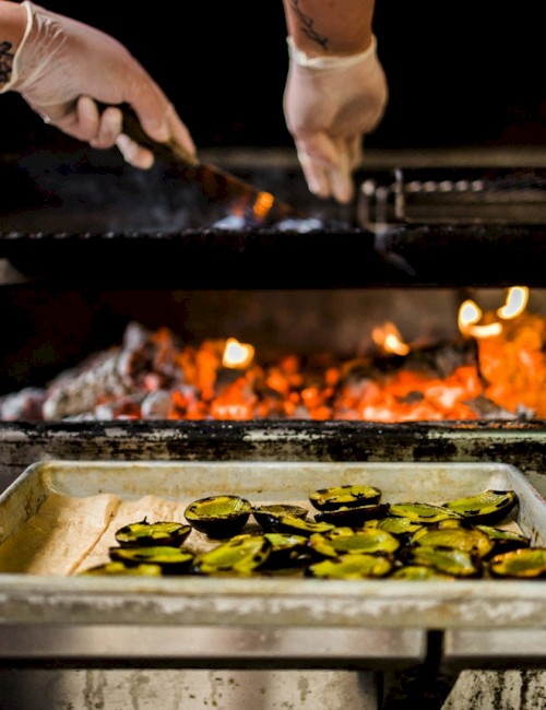 A person wearing gloves is grilling food over an open flame while vegetables are being roasted in a tray below the grill in this kitchen scene.