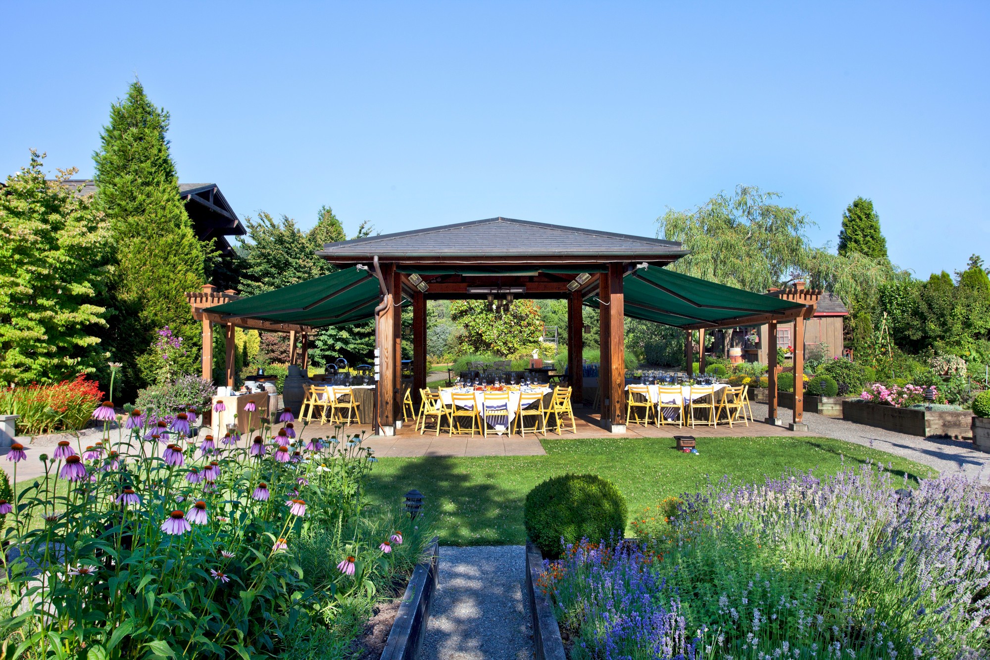 A garden pergola structure with green canopy shades, surrounded by plants and flowers, with tables and yellow chairs set up underneath.