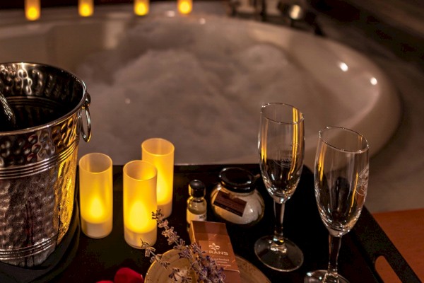 The image shows a romantic setup with champagne in an ice bucket, candles, two glasses, and other small items in front of a bathtub filled with bubbles.