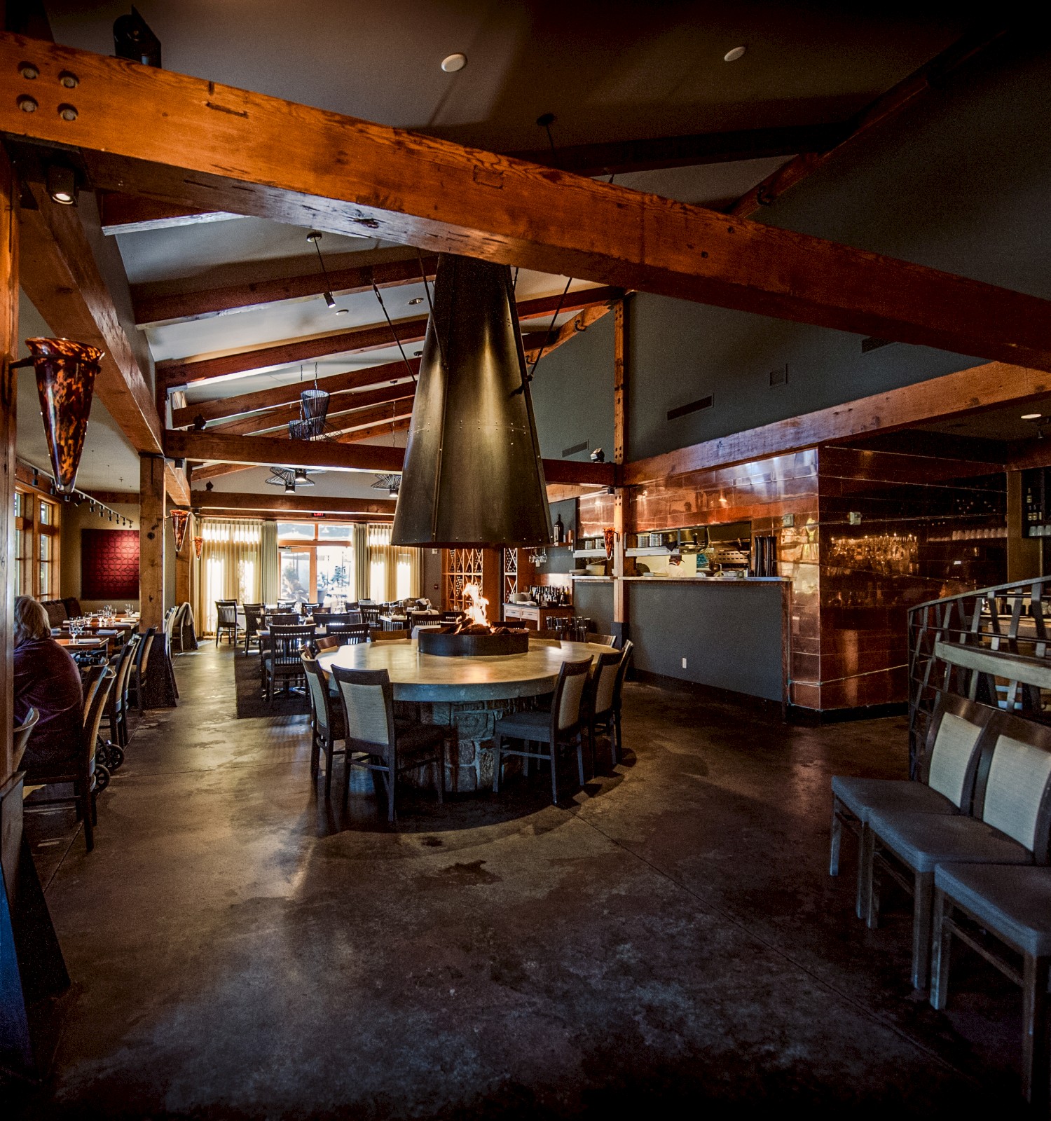 A modern, warmly lit restaurant with wooden beams, a central round table, a bar counter with stools, and seating areas along the walls.