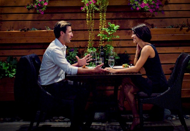 A couple is seated at an outdoor restaurant, engaging in conversation with drinks on the table, surrounded by plants and wooden decor.