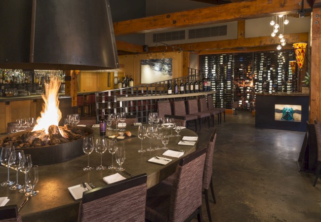 The image shows a cozy restaurant interior with a round table set with glasses and napkins, a central fire pit, and a wine display in the background.