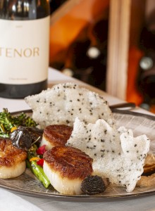 The image shows a gourmet dish with seared scallops, broccolini, and crispy rice paper, accompanied by a glass of white wine and a bottle labeled 