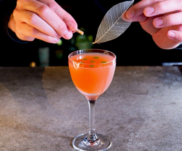 A person is garnishing a pink cocktail with a decorative leaf on a bar counter, creating an artistic presentation.