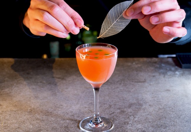 A person is garnishing a pink cocktail with a decorative leaf on a bar counter, creating an artistic presentation.