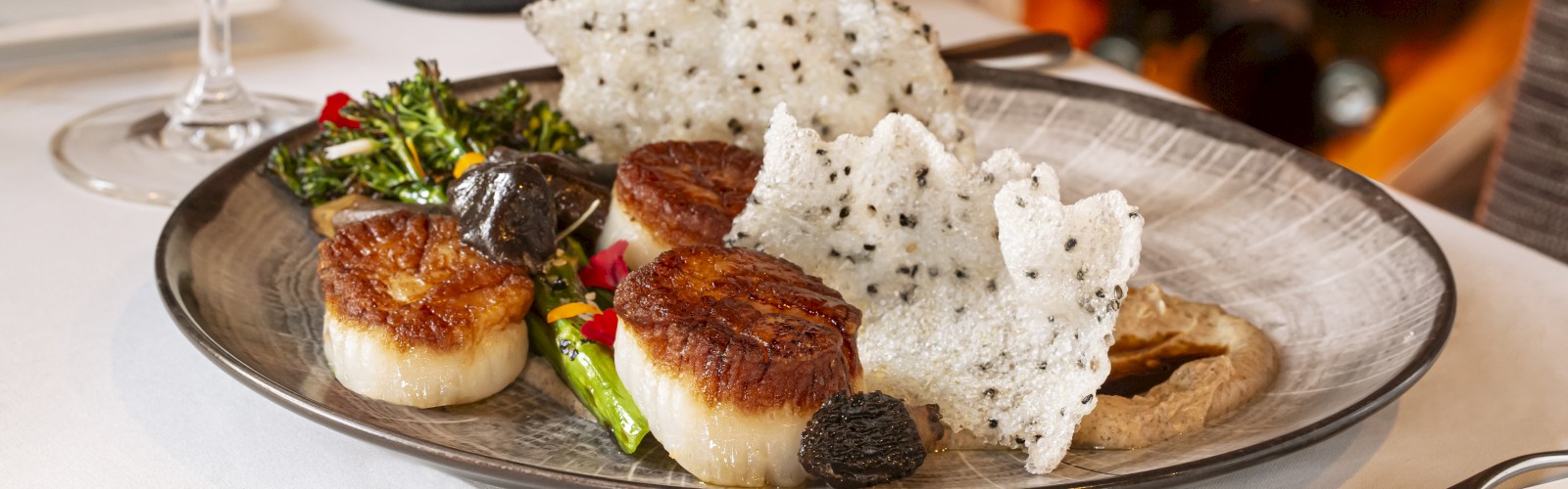 The image shows a gourmet dish with scallops, vegetables, truffle, and rice crackers, accompanied by a glass of white wine and a bottle labeled