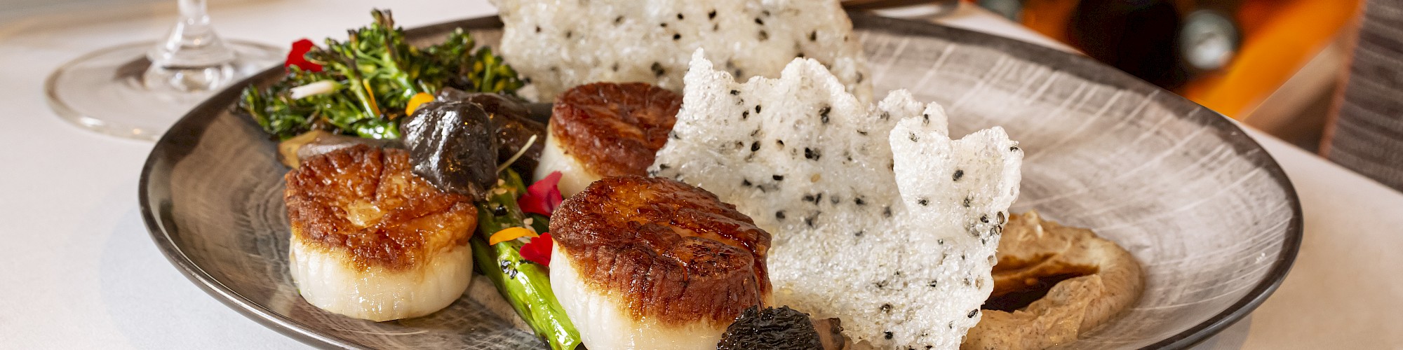 The image shows a gourmet dish with scallops, vegetables, truffle, and rice crackers, accompanied by a glass of white wine and a bottle labeled 