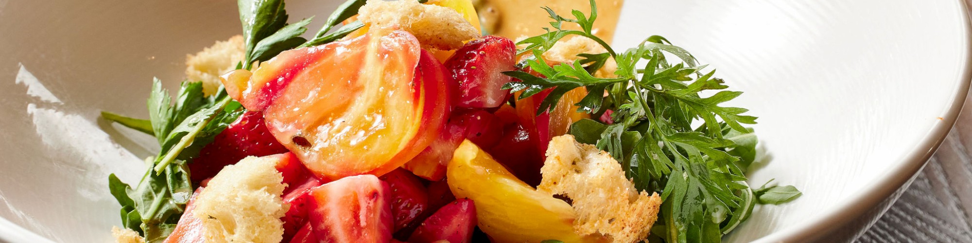 A bowl of fresh salad with tomatoes, greens, croutons, and a creamy dressing on the side, garnished with herbs, placed on a wooden surface.