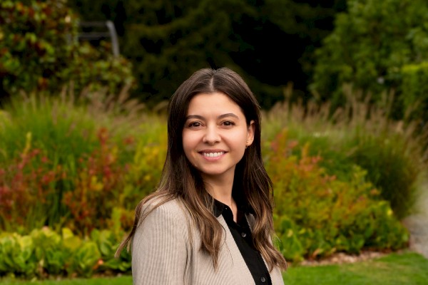 A woman is smiling in an outdoor setting, surrounded by green and colorful plants, with a blurred background of trees and foliage.