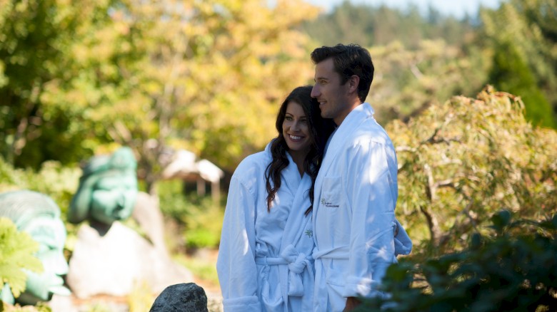 A couple in white robes smiles while standing outdoors in a lush garden. They appear relaxed, enjoying the beautiful scenery around them.