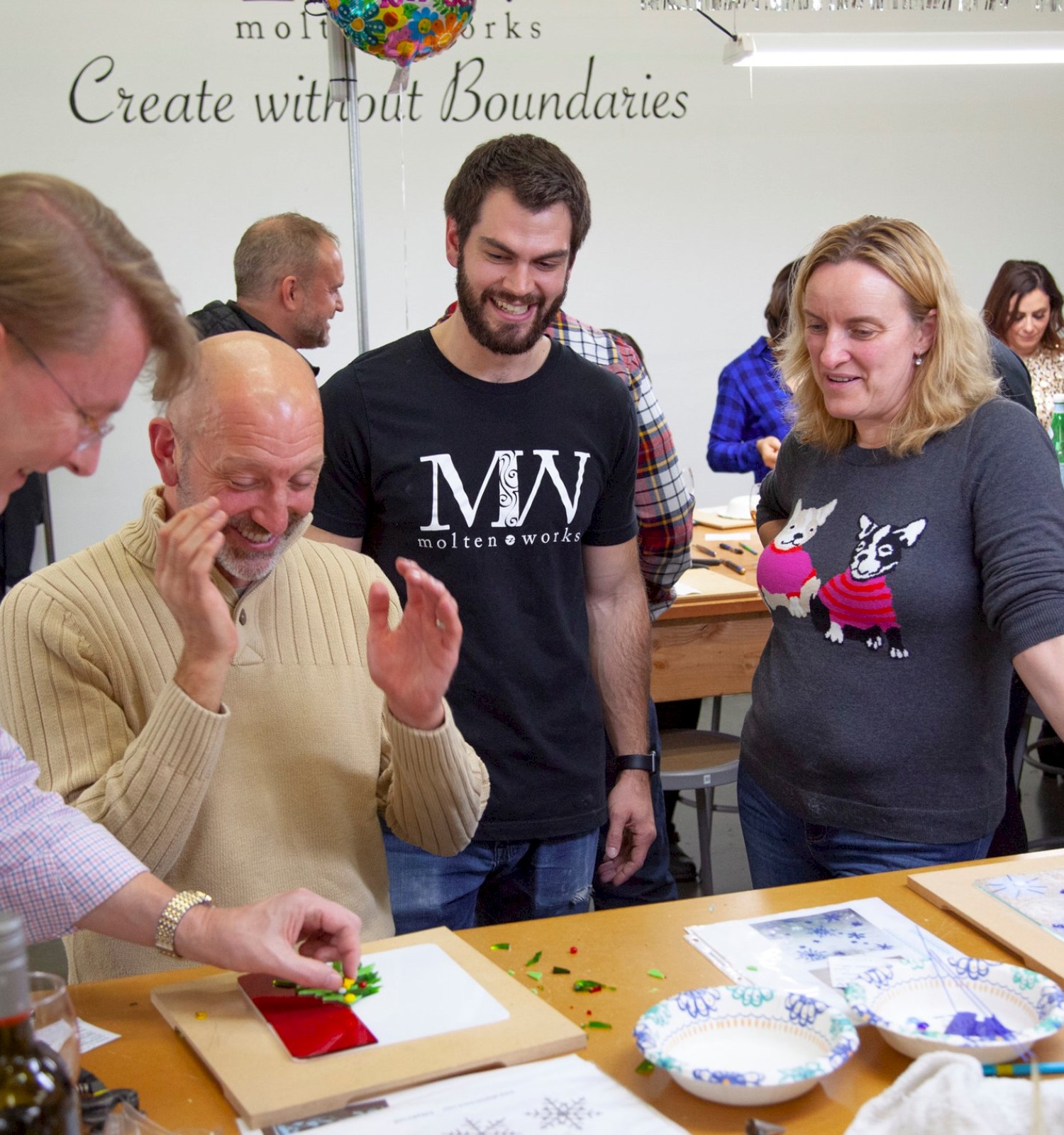 A group of people are gathered around a table, engaging in a crafts activity, with one person appearing to demonstrate something.