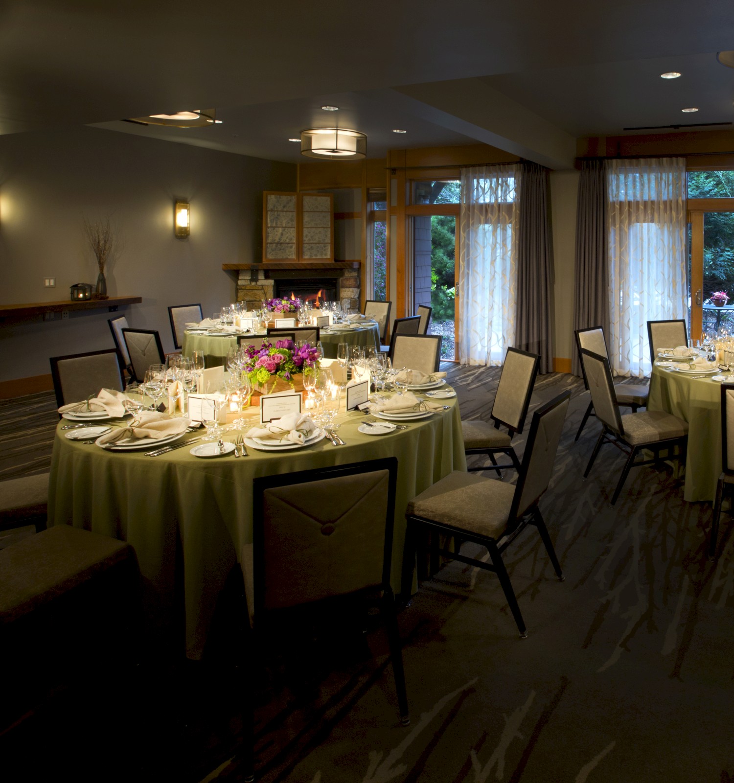 A dining room with round tables set for a formal event, featuring white tablecloths, glasses, cutlery, and floral centerpieces by large windows.