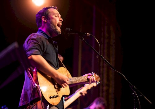 A person is passionately playing a guitar and singing into a microphone on a stage under warm lighting.