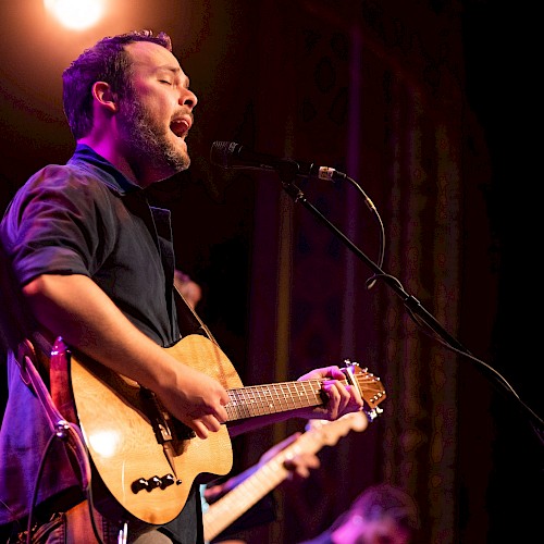 A person is passionately playing a guitar and singing into a microphone on a stage under warm lighting.