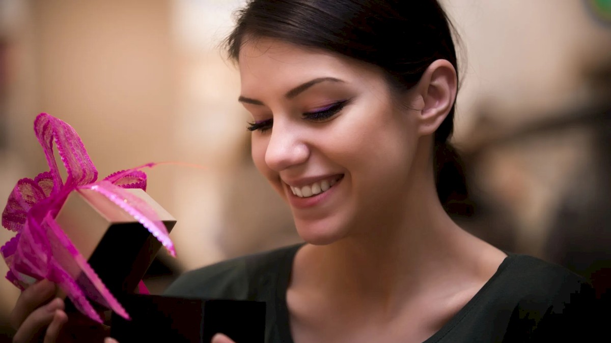 A person is smiling while opening a small black gift box with a pink ribbon, appearing delighted with its contents.