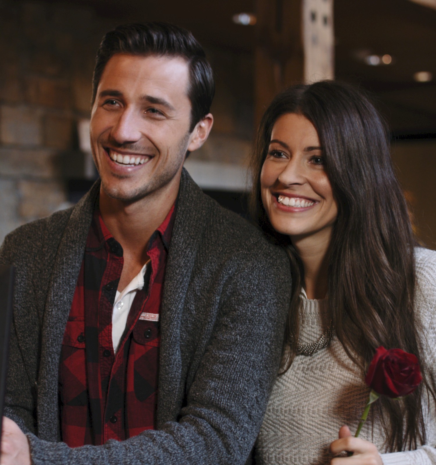 Two people smile together indoors, one holding a single red rose, standing near a counter.