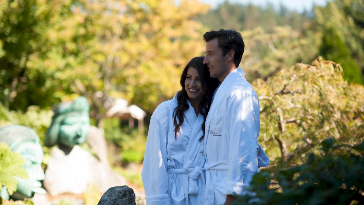 A couple in white bathrobes stands together in a garden with lush greenery and scenic background, enjoying a sunny day together.