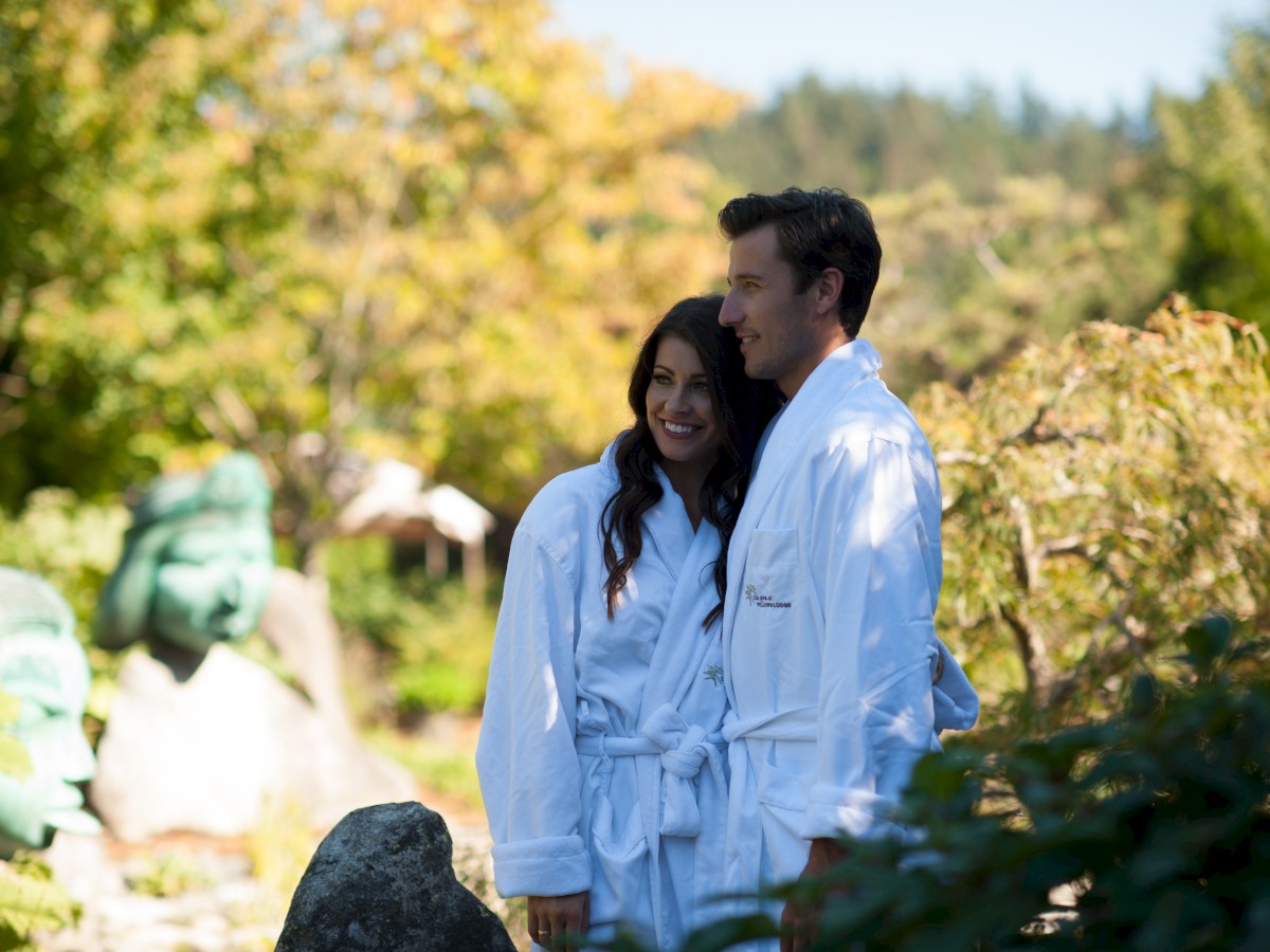 A couple in white bathrobes stands together in a garden with lush greenery and scenic background, enjoying a sunny day together.