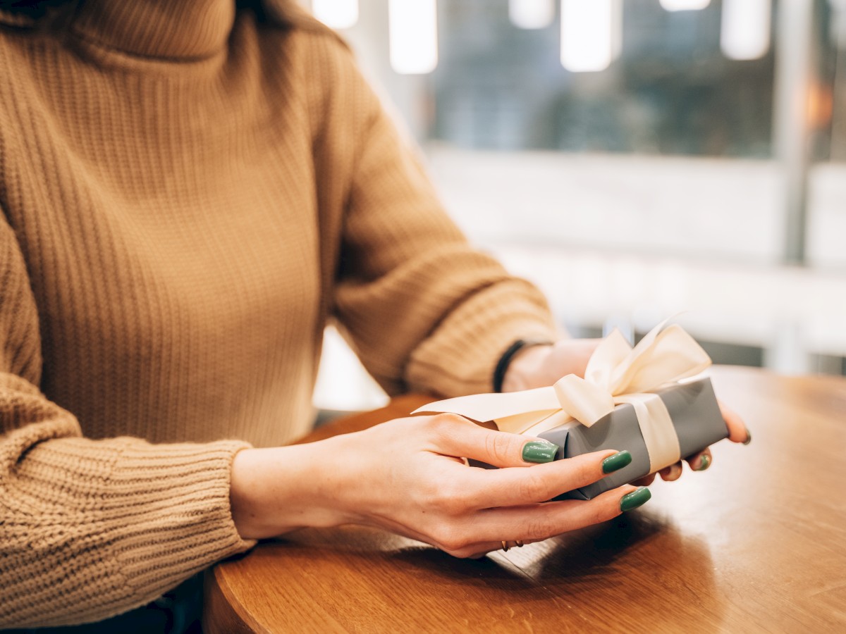 A person wearing a brown sweater holds a wrapped gift box with a bow, sitting at a wooden table.