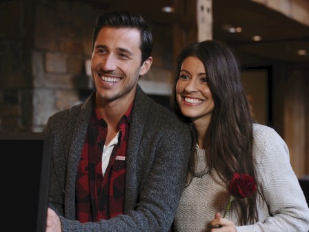 A smiling couple stands together, with the woman holding a single red rose, inside a cozy, rustic lodge with wooden beams and stone walls.