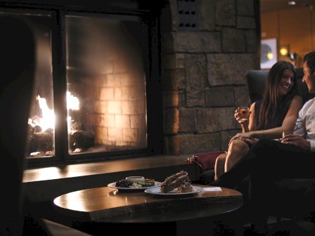 A couple enjoys a cozy moment by a fireplace, sharing drinks and food, with soft lighting creating a warm and romantic ambiance in the room.