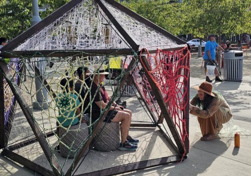 A geometric structure made of metal and yarn with people sitting inside while another person on the outside interacts with it in a public area.