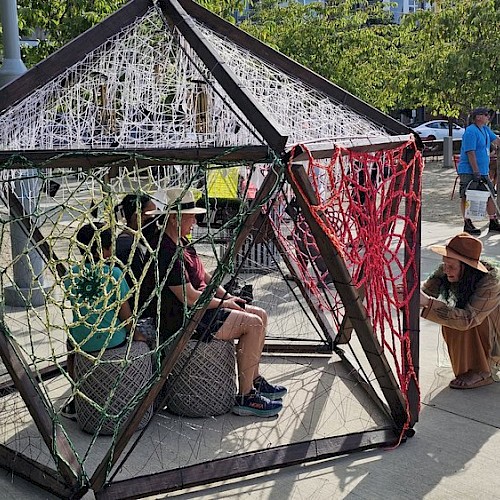 A geometric structure made of metal and yarn with people sitting inside while another person on the outside interacts with it in a public area.