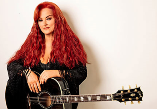 A woman with long red hair is standing against a white background, holding a black acoustic guitar with gold tuning pegs, and wearing a black top.