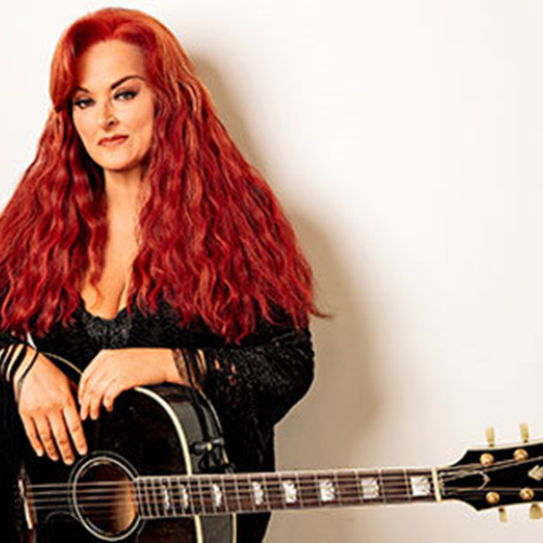 A woman with long red hair is standing against a white background, holding a black acoustic guitar with gold tuning pegs, and wearing a black top.