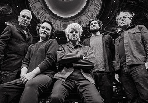 Five men are posed in a black and white photo with a decorative ceiling above them, creating a dramatic and symmetrical composition.