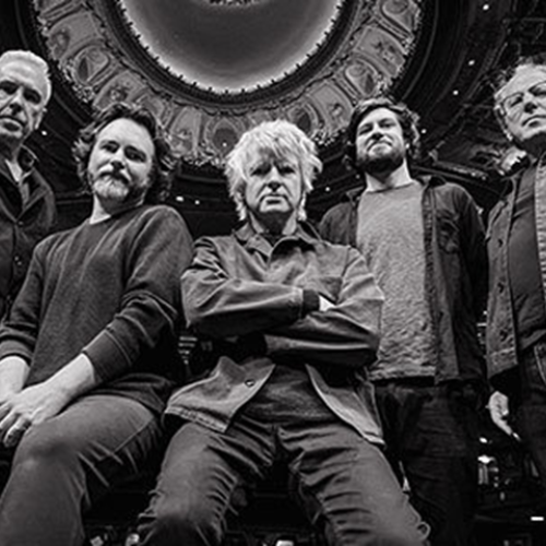 Five men are posed in a black and white photo with a decorative ceiling above them, creating a dramatic and symmetrical composition.