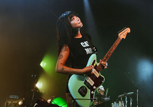 A person with a guitar is performing on stage under colorful lights, wearing a black shirt. Drums and other stage equipment are visible in the background.