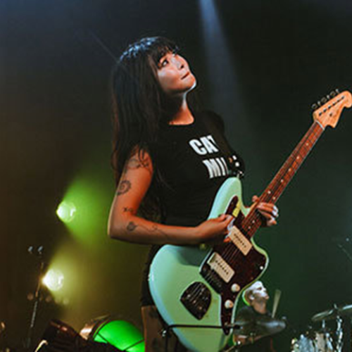 A person with a guitar is performing on stage under colorful lights, wearing a black shirt. Drums and other stage equipment are visible in the background.