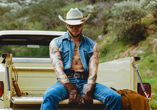 A person in a cowboy hat and denim outfit sits on the tailgate of a vintage pickup truck, surrounded by nature.