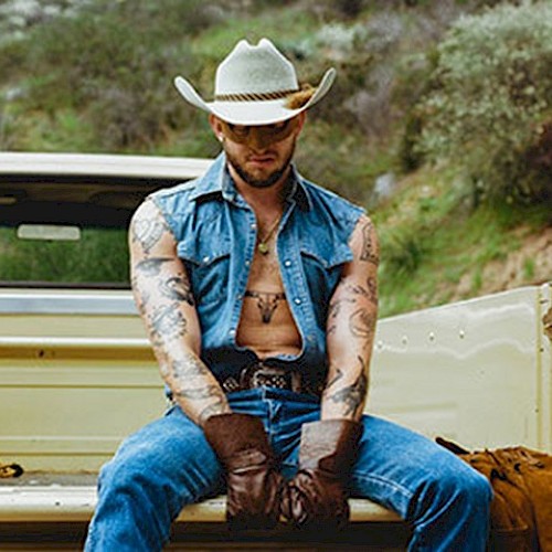 A person in a cowboy hat and denim outfit sits on the tailgate of a vintage pickup truck, surrounded by nature.