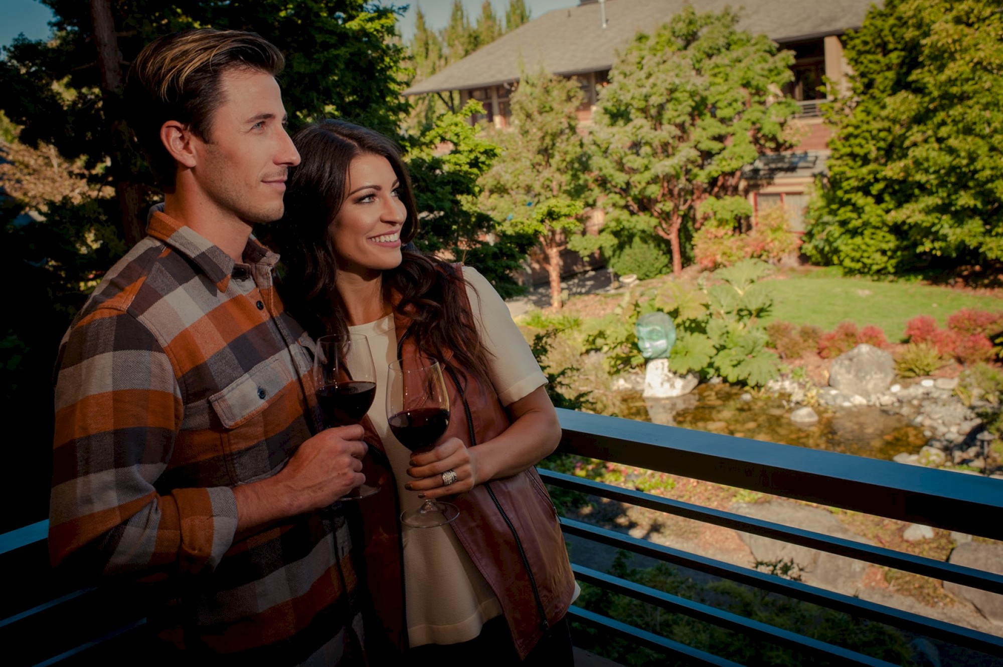 A couple is standing on a balcony, holding wine glasses, and enjoying a scenic view with greenery and a building in the background.