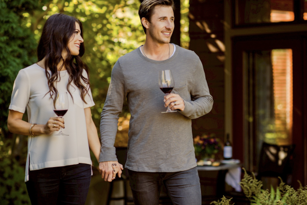 A couple walks outdoors, holding hands and glasses of wine, with leafy greenery and a cozy home patio in the background.