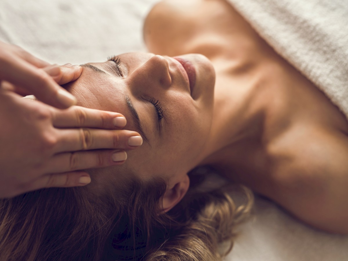 A person is lying down with eyes closed, receiving a head massage.