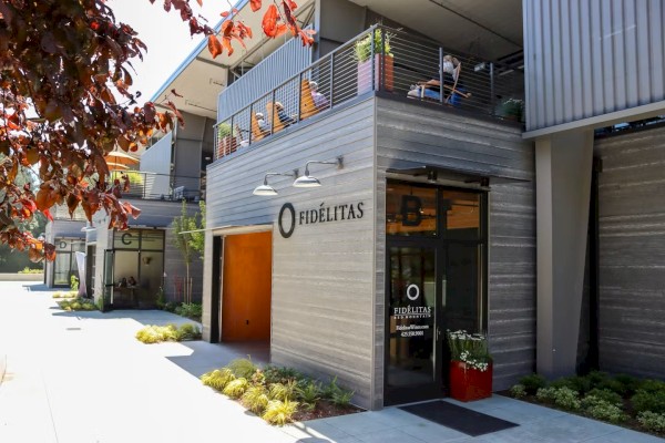 Outdoor scene showing the facade of a modern building with signage “FIDÉLITAS” and “TASTING ROOM open daily,” alongside greenery and a tree.