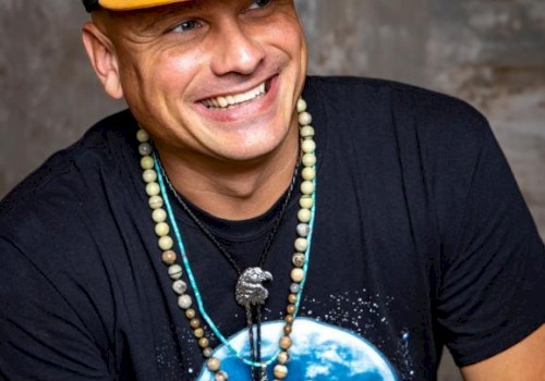 A smiling person is wearing a black cap with a yellow letter, a t-shirt with a moon and wolf design, and multiple necklaces, in front of a grey background.