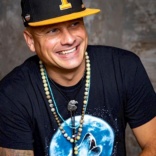 A smiling person is wearing a black cap with a yellow letter, a t-shirt with a moon and wolf design, and multiple necklaces, in front of a grey background.