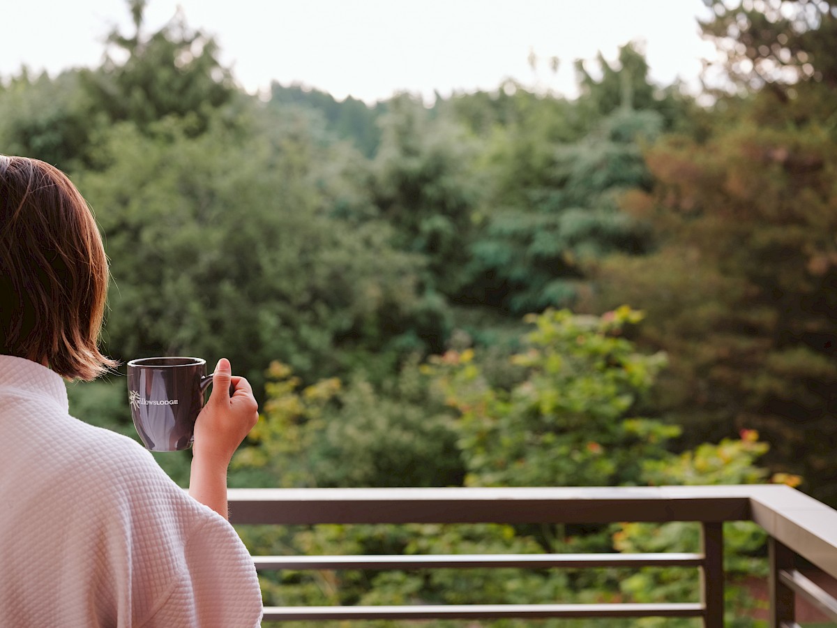 A person in a white robe is holding a mug while standing on a balcony, overlooking a lush, green, forested area in the distance.