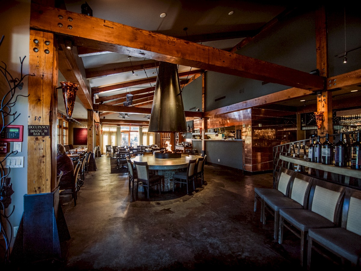 This image shows the interior of a rustic restaurant with wooden beams, a central round table, and chairs lining the walls, creating a cozy ambiance.