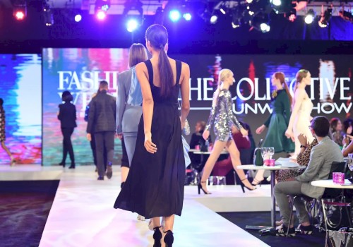 Models are walking down a runway during a fashion show with an audience seated on the sides. The background displays 